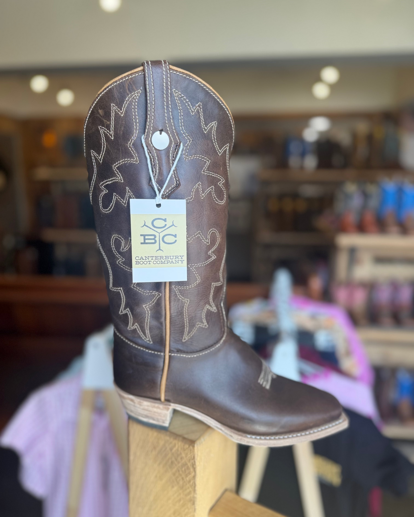 Brown cowboy boot with a visible brand tag on a wooden stool in a store setting.