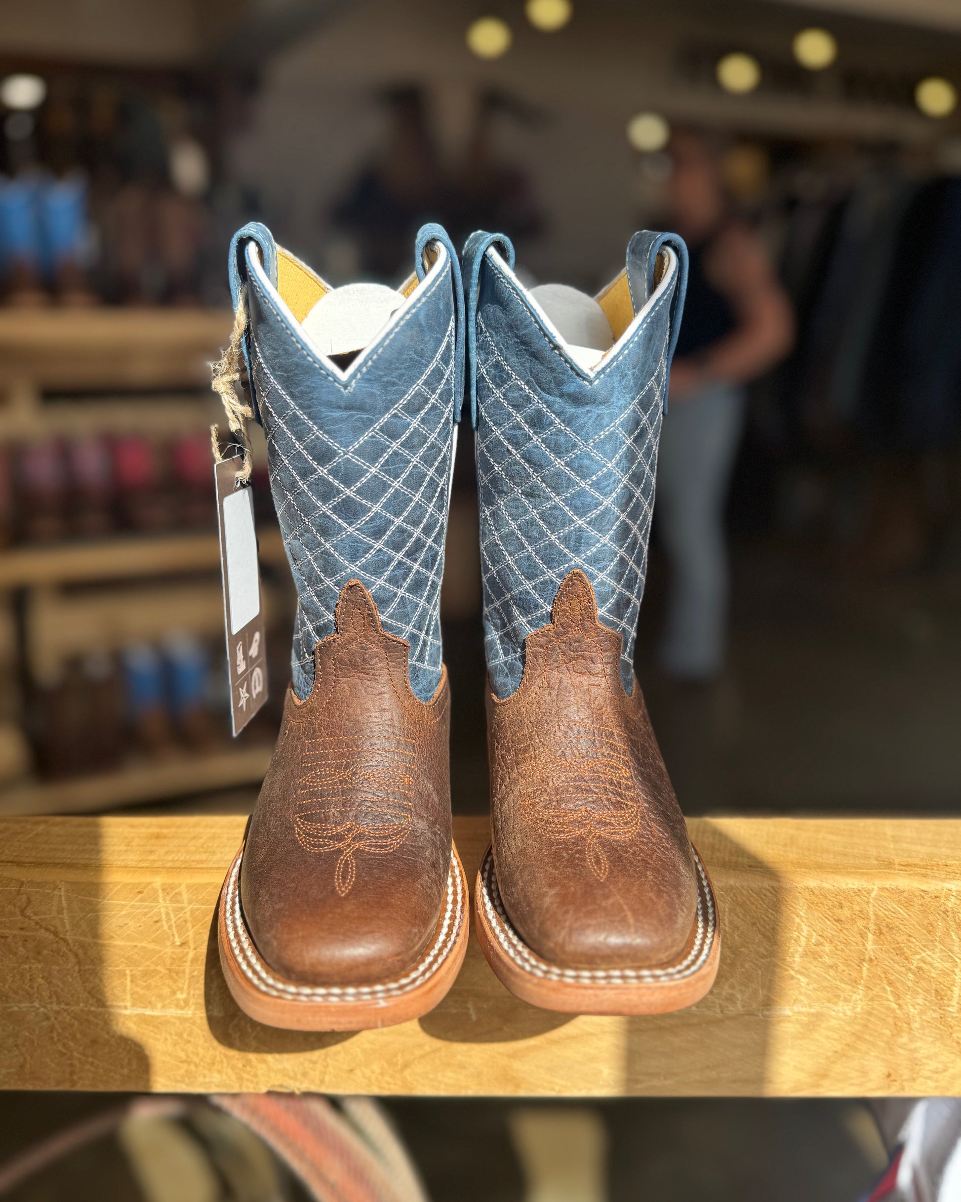 Pair of brown and blue cowboy boots on a wooden surface with a blurred store background.