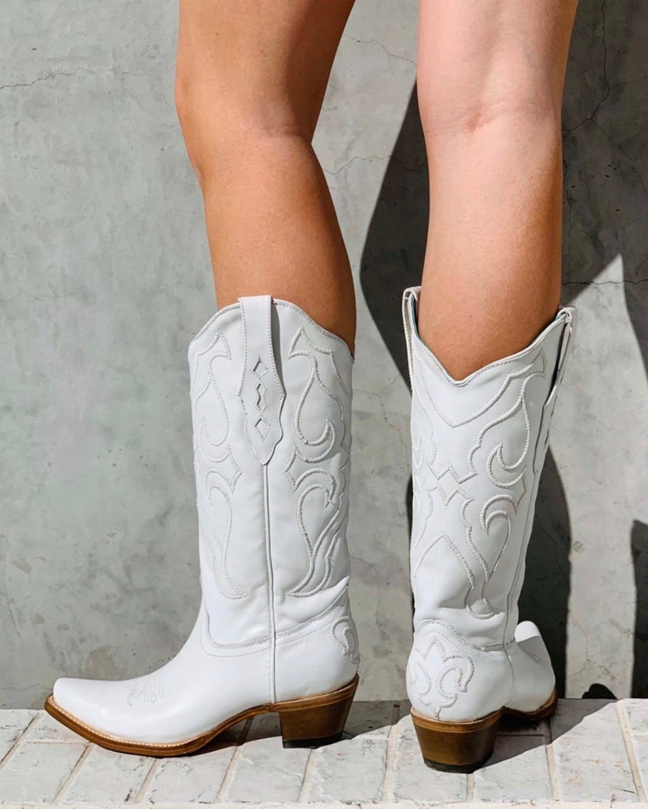White cowboy boots with brown soles worn by a person on a concrete floor.