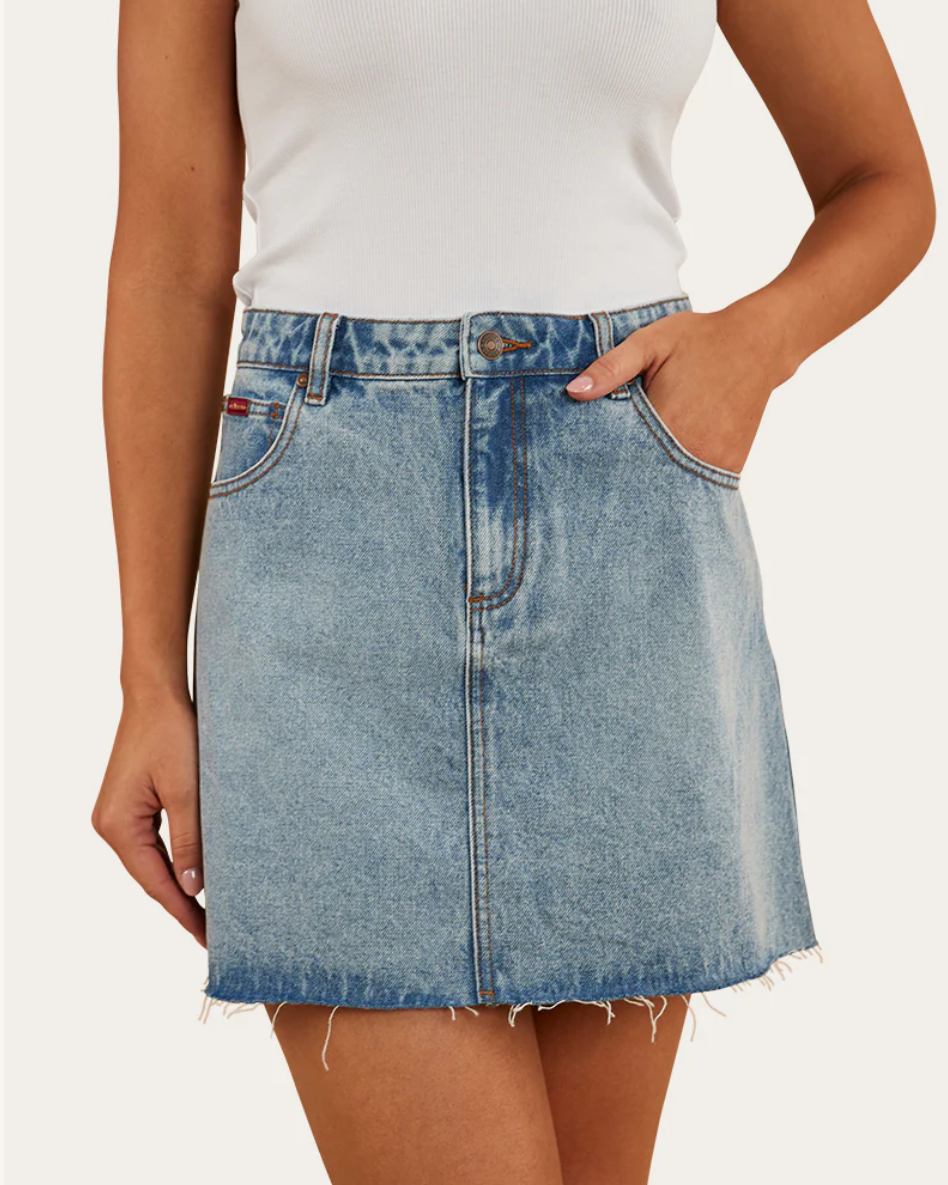 A woman wearing a blue denim skirt with a white top. The skirt reaches mid-thigh and has a frayed hem.