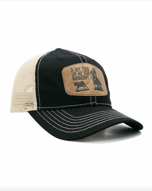 A black and bone trucker-style cap with a curved brim and contrast stitching. The front panel features a brown leather patch with an engraved western-style design, showing a cowboy tying up a calf. The patch includes the phrase 'A BIT TIED UP AT THE MOMENT' in bold capital letters. The cap has a black fabric front and a beige mesh back, offering a breathable and rugged look.