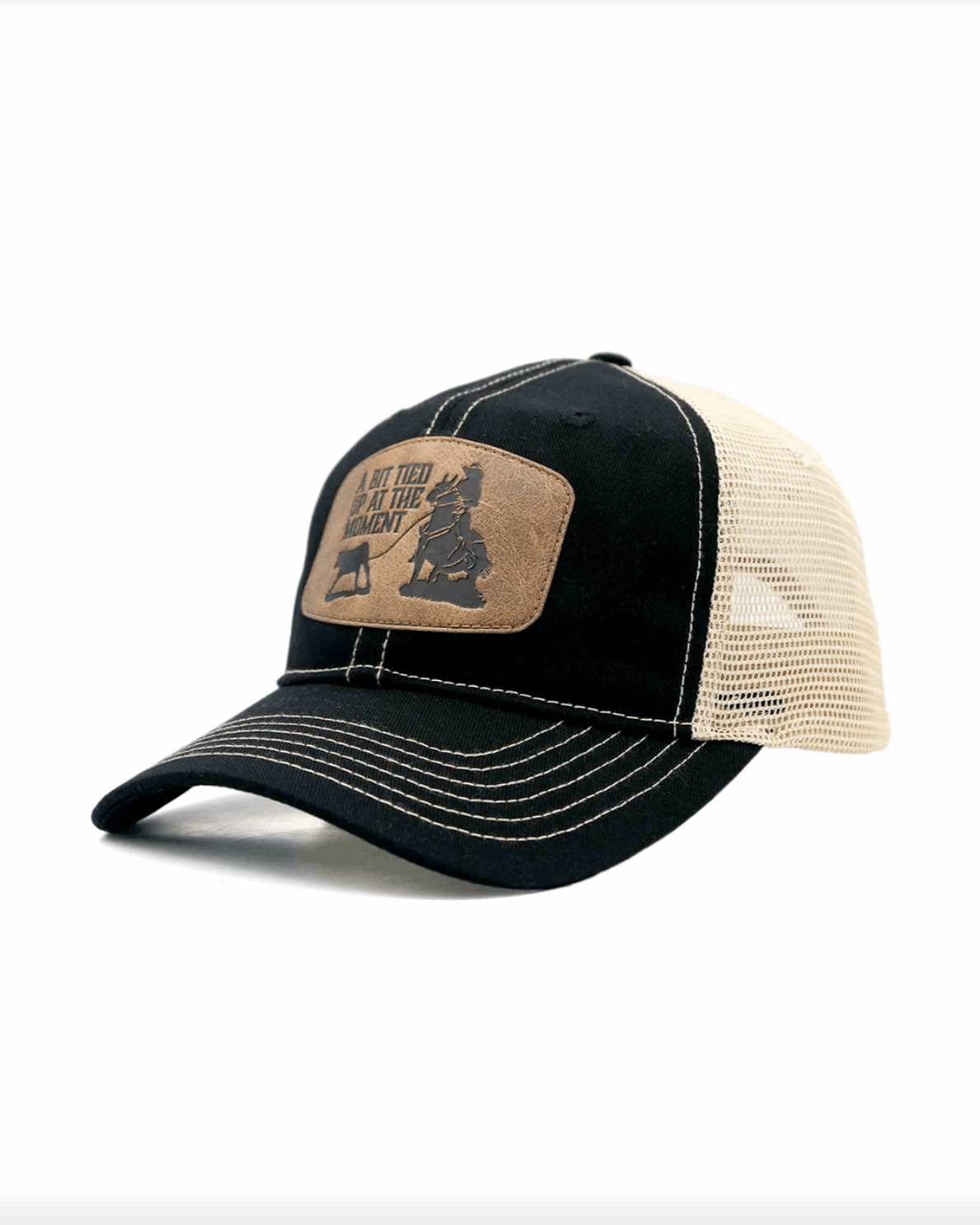 A black and bone trucker-style cap with a curved brim and contrast stitching. The front panel features a brown leather patch with an engraved western-style design, showing a cowboy tying up a calf. The patch includes the phrase 'A BIT TIED UP AT THE MOMENT' in bold capital letters. The cap has a black fabric front and a beige mesh back, offering a breathable and rugged look.