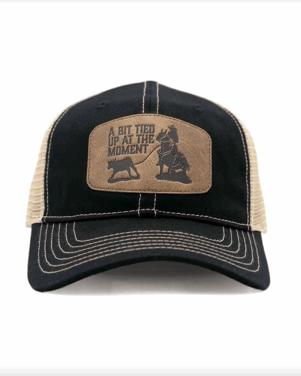 A black and bone trucker-style cap with a curved brim and contrast stitching. The front panel features a brown leather patch with an engraved western-style design, showing a cowboy tying up a calf. The patch includes the phrase 'A BIT TIED UP AT THE MOMENT' in bold capital letters. The cap has a black fabric front and a beige mesh back, offering a breathable and rugged look.