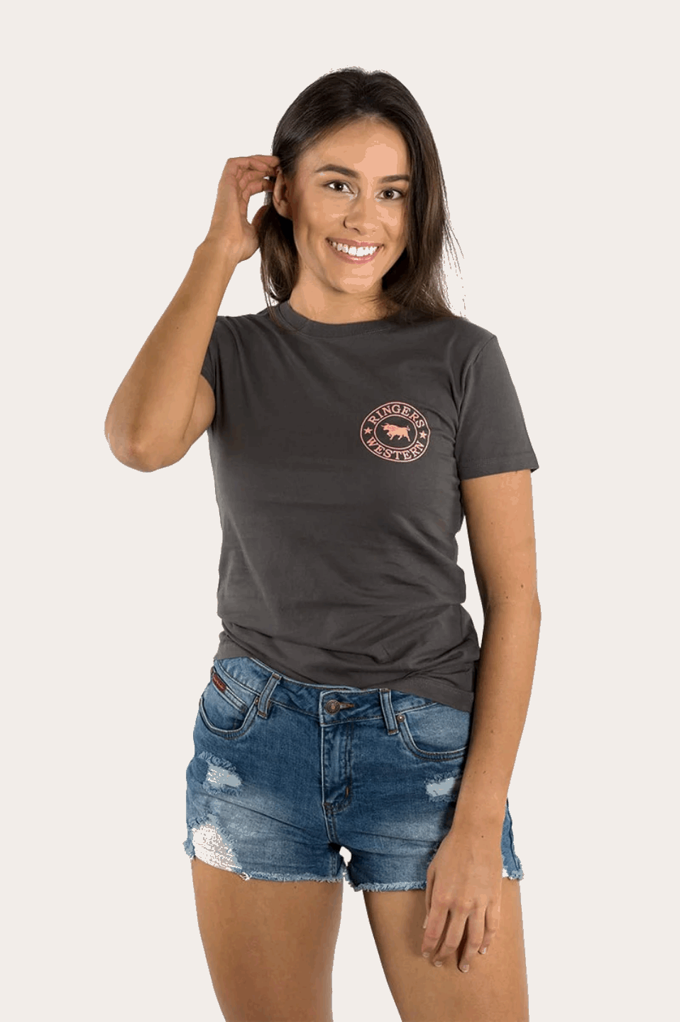 A women's fitted charcoal grey t-shirt featuring a small pink "Ringers Western" logo on the left chest and a large matching logo on the back, showcasing a bull silhouette encircled by stars. The tee has a classic crew neck and short sleeves, styled with denim shorts for a casual, Western-inspired look.