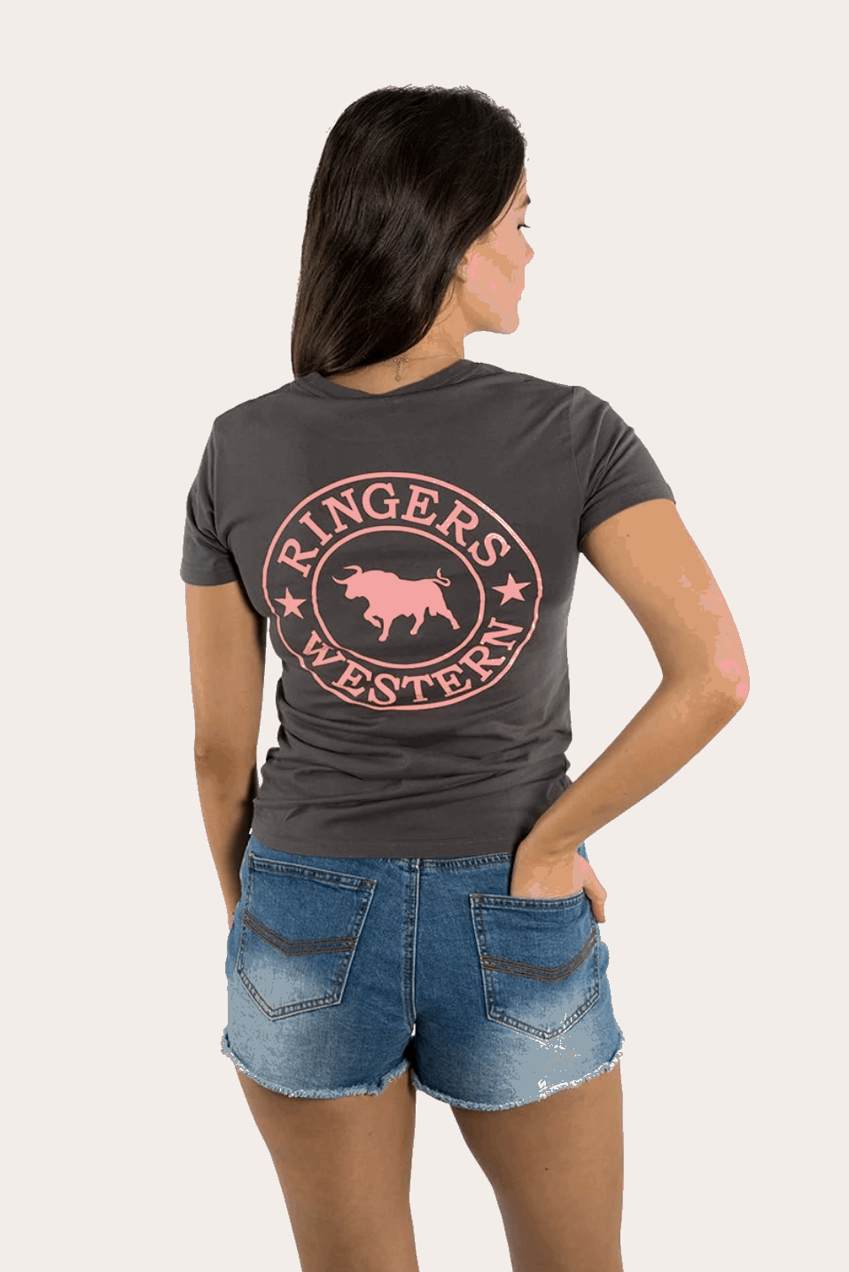 A women's fitted charcoal grey t-shirt featuring a small pink "Ringers Western" logo on the left chest and a large matching logo on the back, showcasing a bull silhouette encircled by stars. The tee has a classic crew neck and short sleeves, styled with denim shorts for a casual, Western-inspired look.