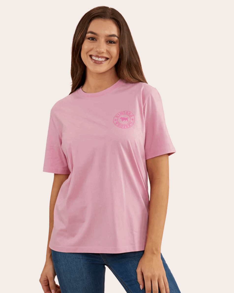 A soft pink, short-sleeved T-shirt with a relaxed fit, featuring a small 'Ringers Western' logo on the left chest area and a larger circular logo with a bull design on the back. The shirt is paired with blue jeans, giving a casual and comfortable look.