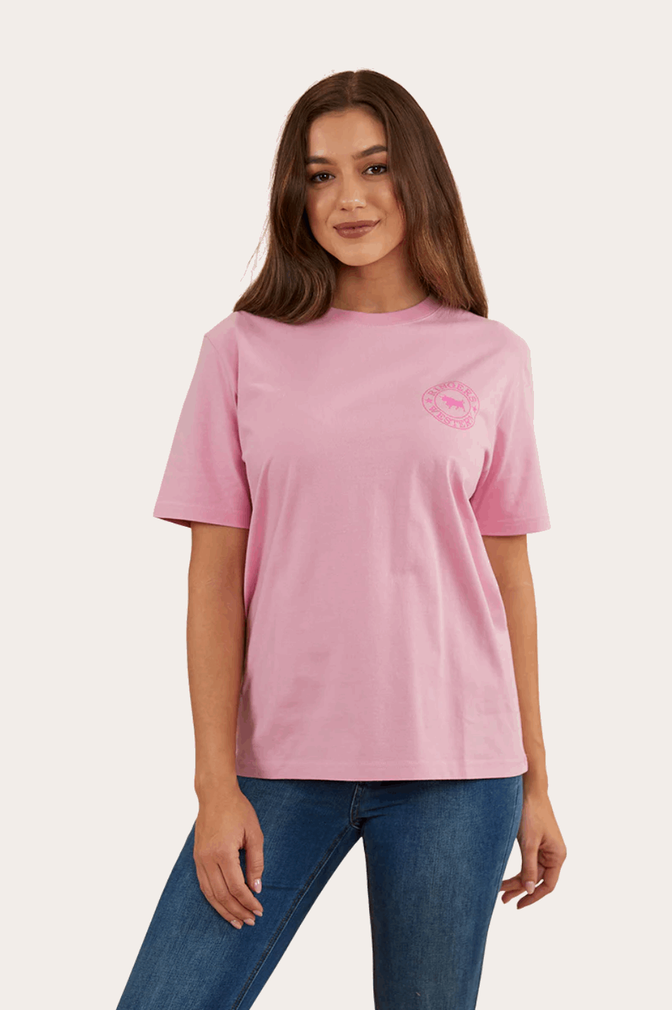 A soft pink, short-sleeved T-shirt with a relaxed fit, featuring a small 'Ringers Western' logo on the left chest area and a larger circular logo with a bull design on the back. The shirt is paired with blue jeans, giving a casual and comfortable look.