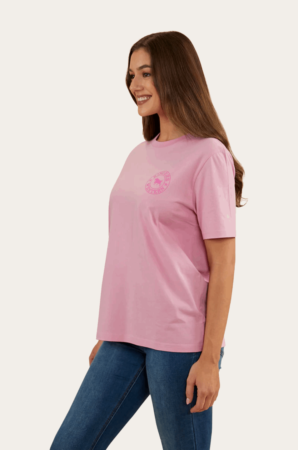 A soft pink, short-sleeved T-shirt with a relaxed fit, featuring a small 'Ringers Western' logo on the left chest area and a larger circular logo with a bull design on the back. The shirt is paired with blue jeans, giving a casual and comfortable look.