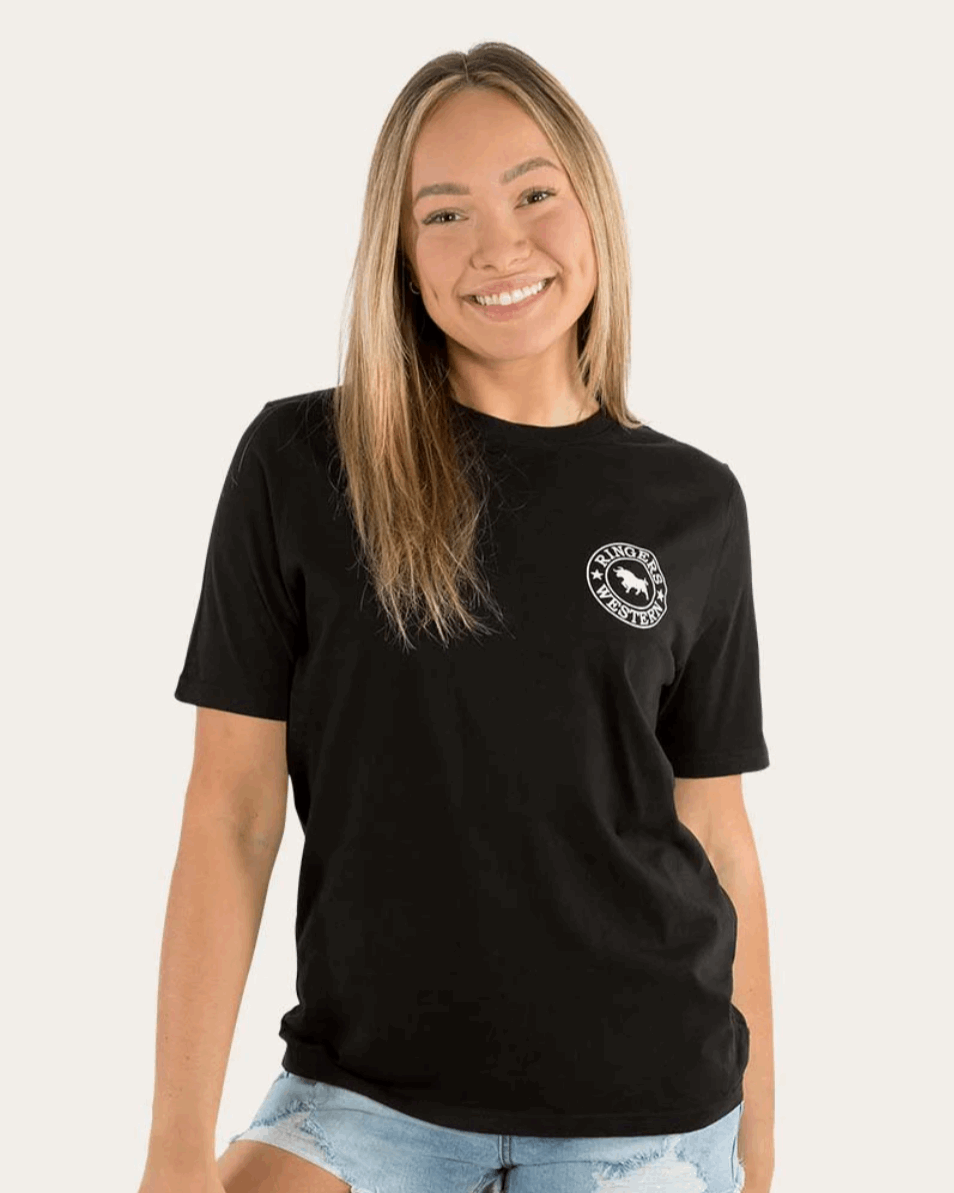 A woman wearing a black Ringers Western t-shirt featuring a small white circular logo on the front left chest and a large white bull logo with text on the back. The tee is paired with distressed denim shorts, creating a casual yet stylish western-inspired look.