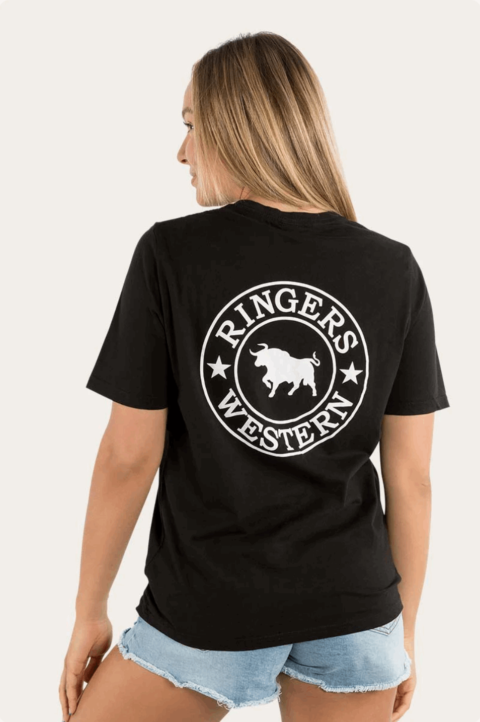 A woman wearing a black Ringers Western t-shirt featuring a small white circular logo on the front left chest and a large white bull logo with text on the back. The tee is paired with distressed denim shorts, creating a casual yet stylish western-inspired look.