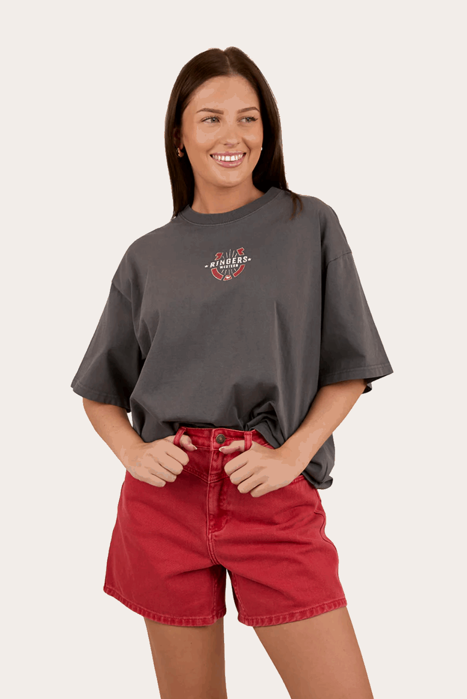 A charcoal gray oversized T-shirt with a small western-inspired "Ringers Western" logo on the front, featuring a horseshoe and arrow design in red, white, and black. The shirt has a relaxed fit and is styled with a light-wash denim skirt. The background includes a rustic, weathered metal wall with brown and silver tones.