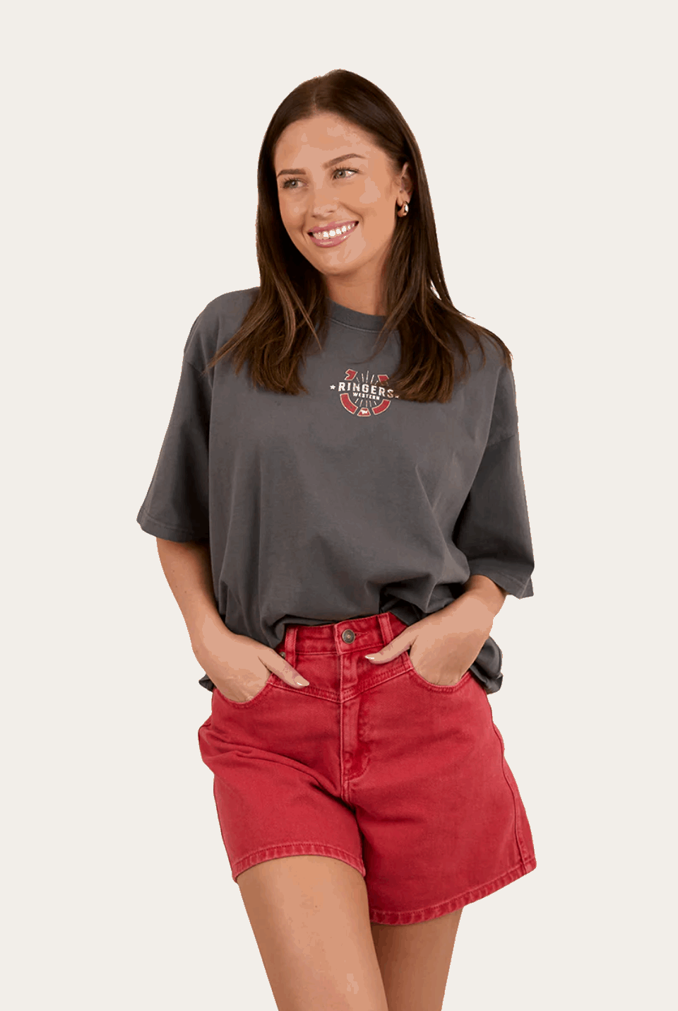 A charcoal gray oversized T-shirt with a small western-inspired "Ringers Western" logo on the front, featuring a horseshoe and arrow design in red, white, and black. The shirt has a relaxed fit and is styled with a light-wash denim skirt. The background includes a rustic, weathered metal wall with brown and silver tones.