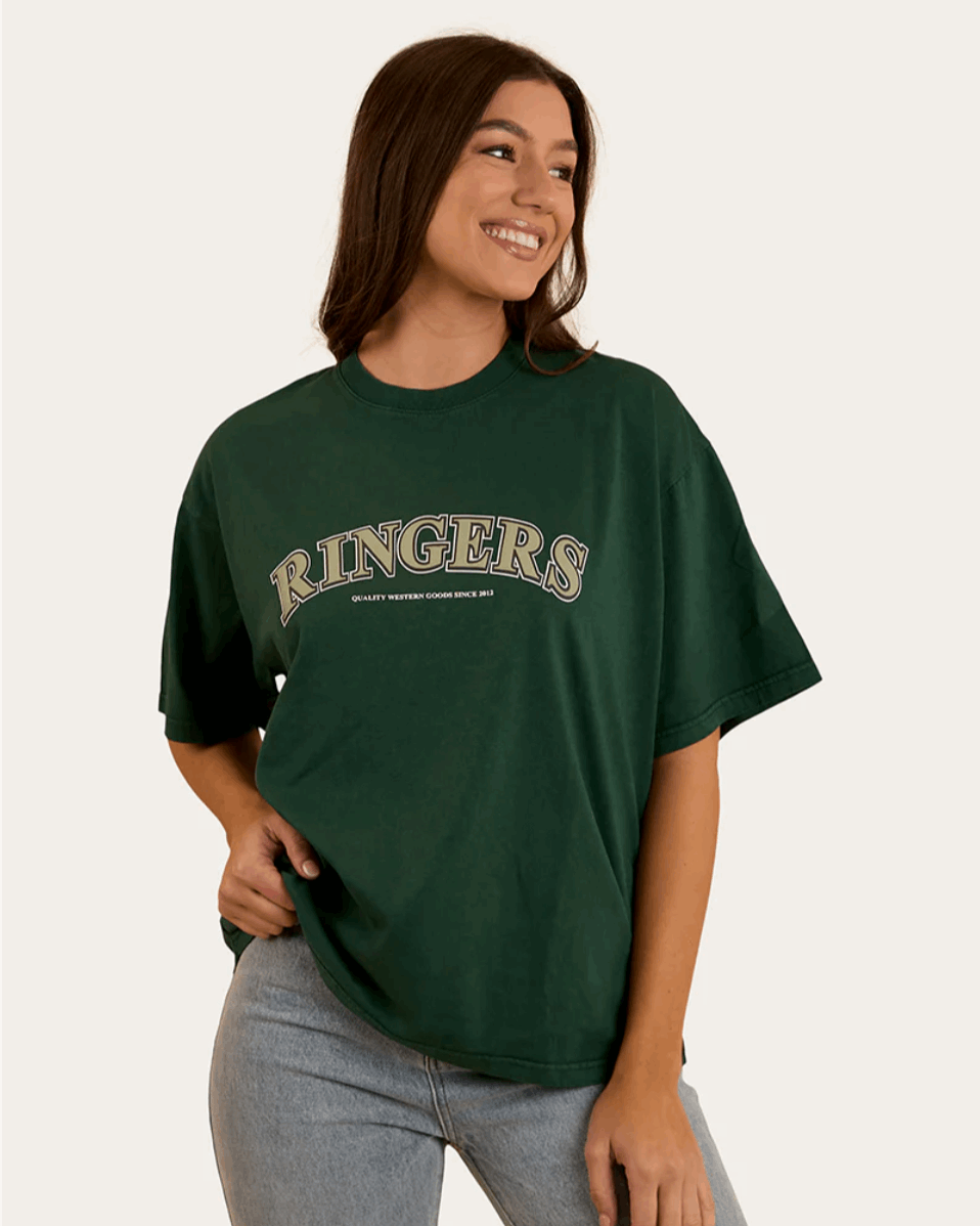 A woman wears a dark green oversized T-shirt with "Ringers" in bold, collegiate-style lettering across the chest. The T-shirt has a relaxed fit with a classic crew neckline and dropped shoulders, offering a casual and comfortable look. She pairs it with light-wash denim jeans, complementing the effortless, laid-back aesthetic. In the back view, the plain green fabric extends smoothly, highlighting the loose fit and soft drape of the material.