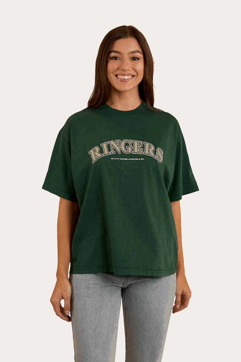 A woman wears a dark green oversized T-shirt with "Ringers" in bold, collegiate-style lettering across the chest. The T-shirt has a relaxed fit with a classic crew neckline and dropped shoulders, offering a casual and comfortable look. She pairs it with light-wash denim jeans, complementing the effortless, laid-back aesthetic. In the back view, the plain green fabric extends smoothly, highlighting the loose fit and soft drape of the material.