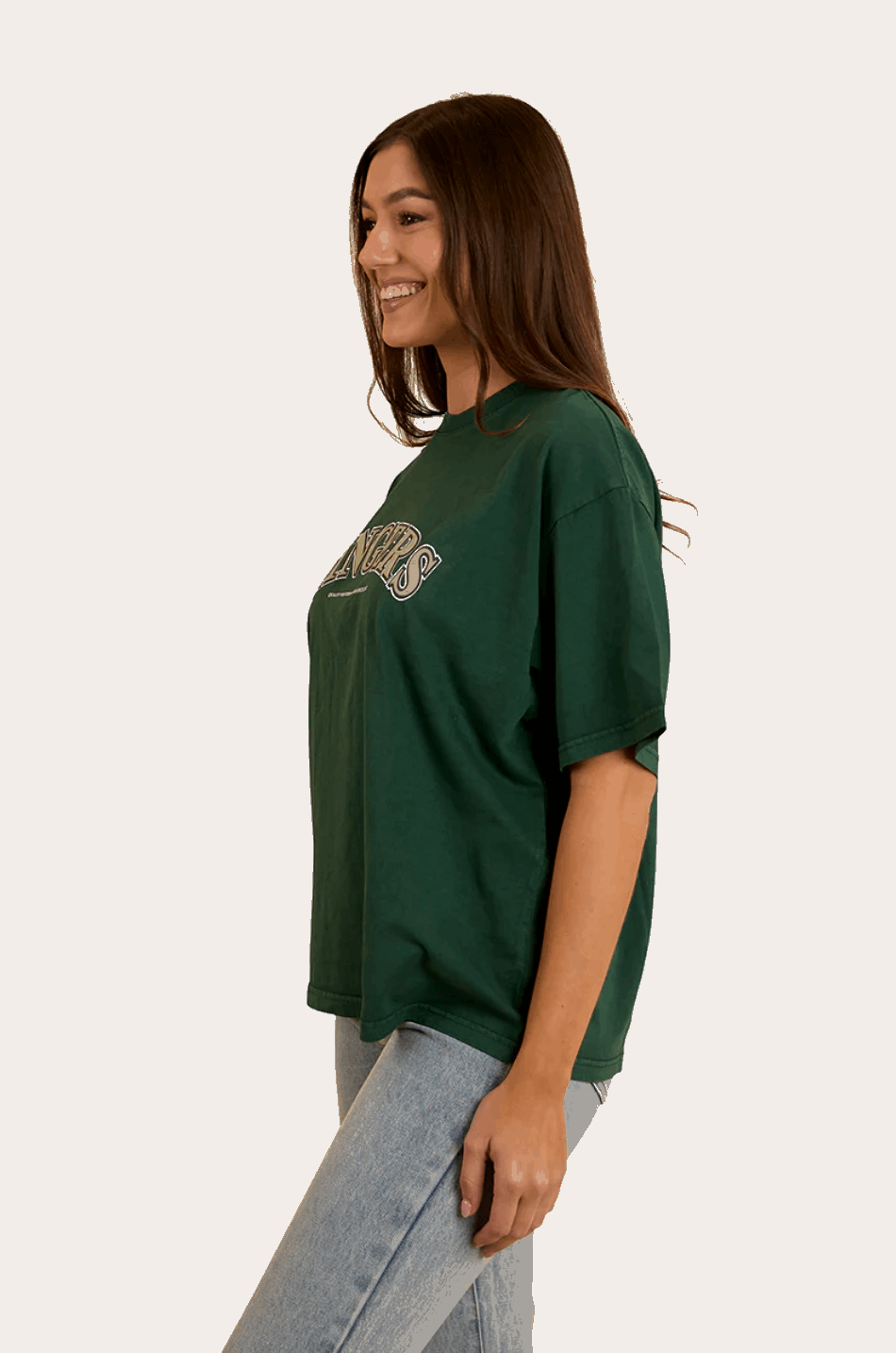 A woman wears a dark green oversized T-shirt with "Ringers" in bold, collegiate-style lettering across the chest. The T-shirt has a relaxed fit with a classic crew neckline and dropped shoulders, offering a casual and comfortable look. She pairs it with light-wash denim jeans, complementing the effortless, laid-back aesthetic. In the back view, the plain green fabric extends smoothly, highlighting the loose fit and soft drape of the material.