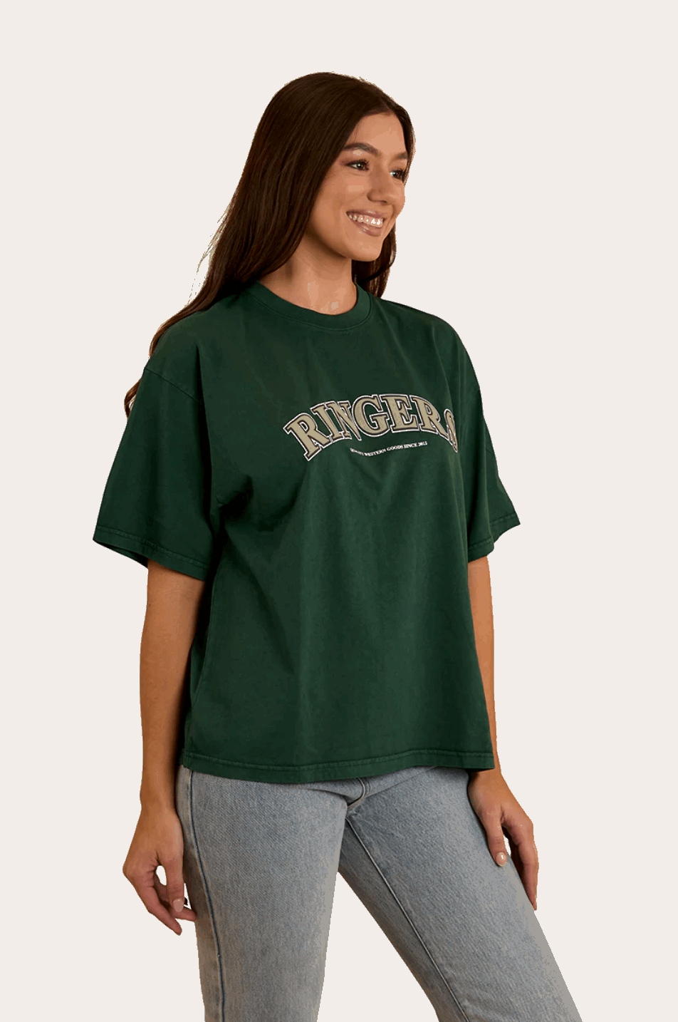 A woman wears a dark green oversized T-shirt with "Ringers" in bold, collegiate-style lettering across the chest. The T-shirt has a relaxed fit with a classic crew neckline and dropped shoulders, offering a casual and comfortable look. She pairs it with light-wash denim jeans, complementing the effortless, laid-back aesthetic. In the back view, the plain green fabric extends smoothly, highlighting the loose fit and soft drape of the material.