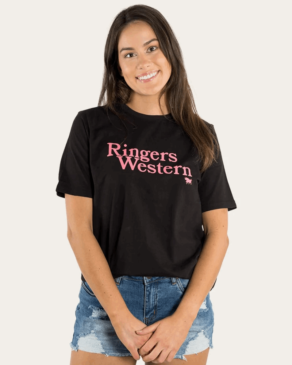 A young woman with long brown hair and a bright smile wears a black Ringers Western t-shirt with pink lettering across the chest. The tee has a relaxed fit and is paired with distressed blue denim shorts. She stands against a neutral background, giving a casual and stylish western-inspired look.