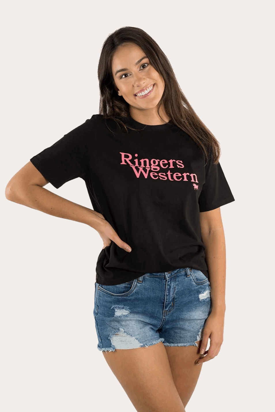 A young woman with long brown hair and a bright smile wears a black Ringers Western t-shirt with pink lettering across the chest. The tee has a relaxed fit and is paired with distressed blue denim shorts. She stands against a neutral background, giving a casual and stylish western-inspired look.