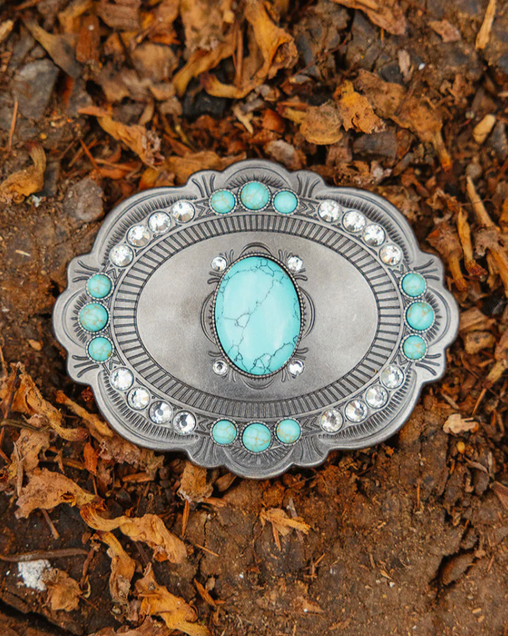 Silver belt buckle with turquoise stone on a bed of soil and leaves