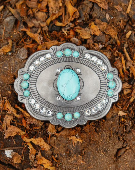 Silver belt buckle with turquoise stone on a bed of soil and leaves