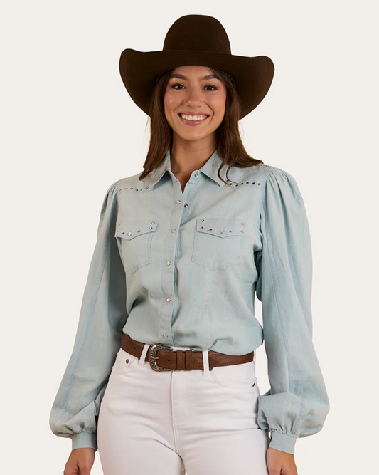 Woman wearing a light blue denim shirt, white pants, and a brown cowboy hat on a white background