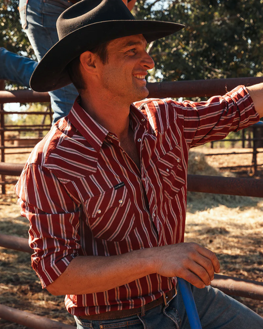 Man wearing a cowboy hat and red striped shirt standing in a rural setting.