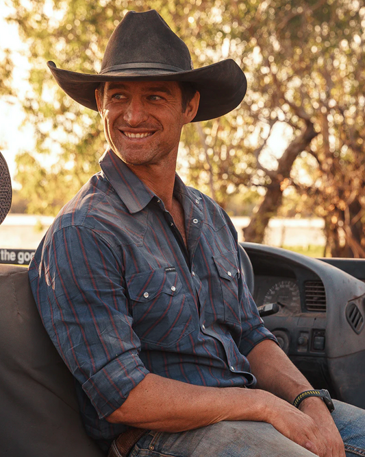 Man wearing a cowboy hat and blue shirt sitting in a vehicle with a blurred natural background