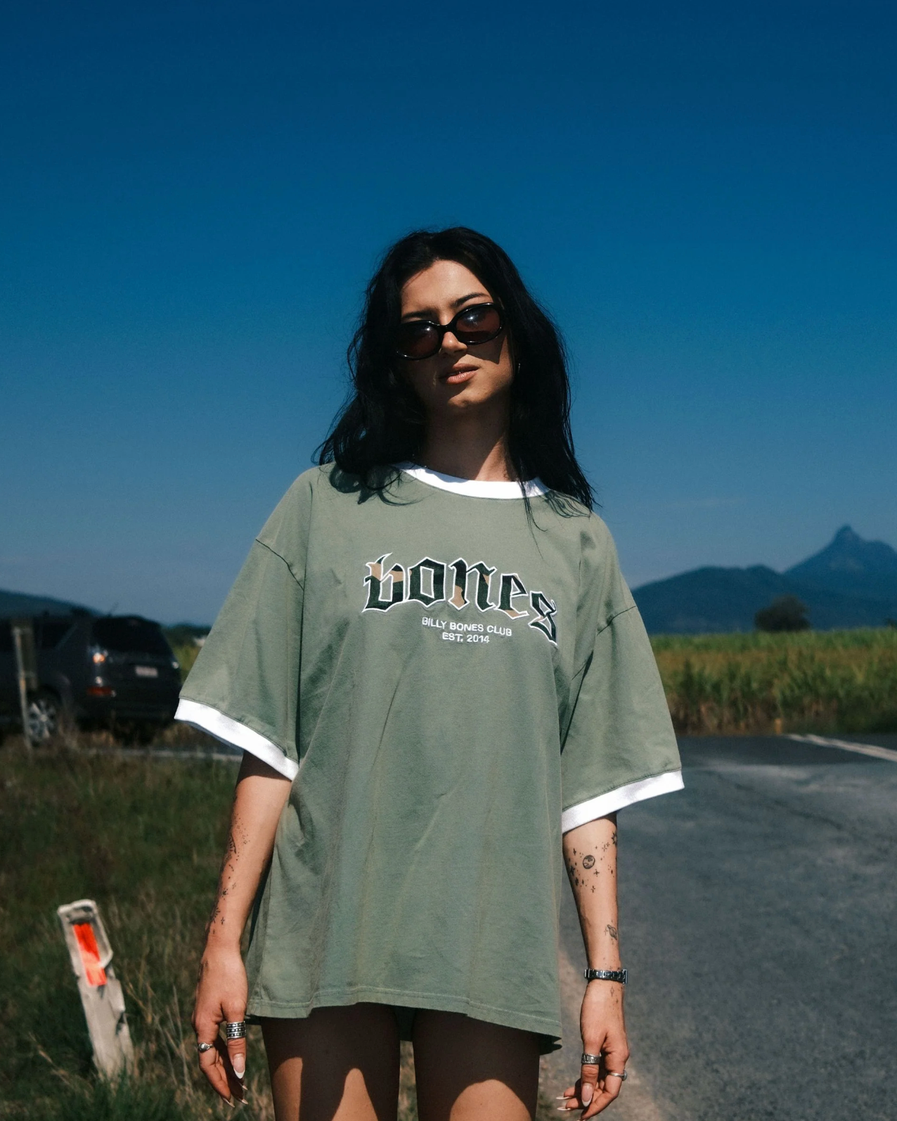 Person wearing a green oversized t-shirt with text, standing on a road with mountains in the background.