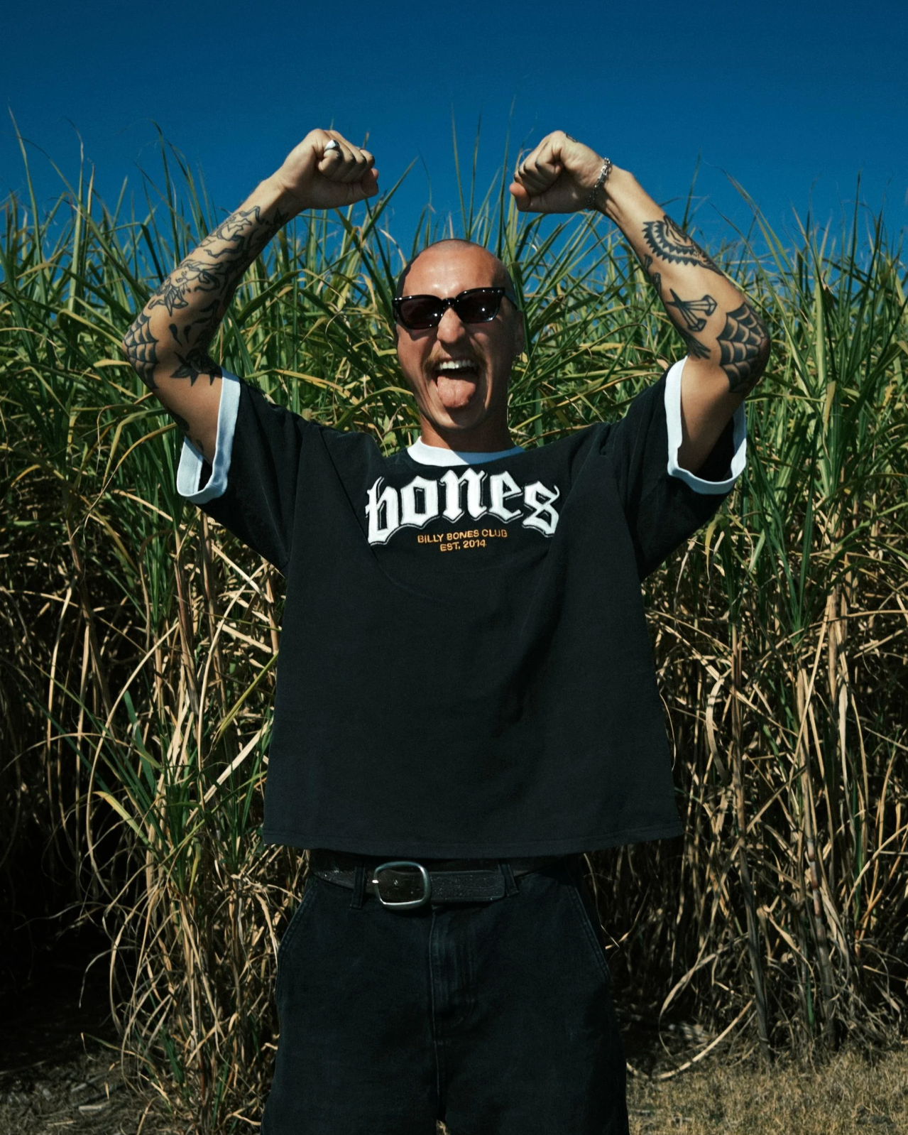 Man in a black 'Bones' t-shirt with arms raised in front of tall grass