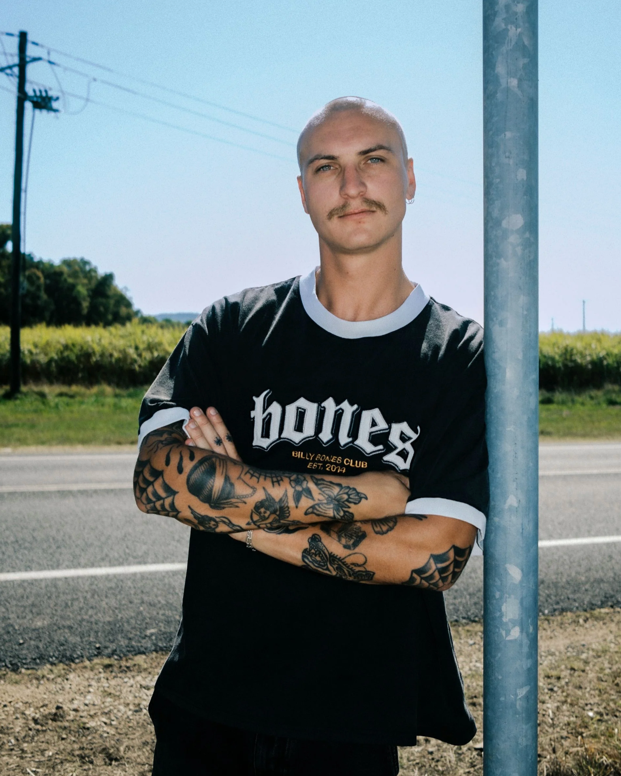 Man wearing a black 'bones' t-shirt with tattoos standing by a road and power pole.