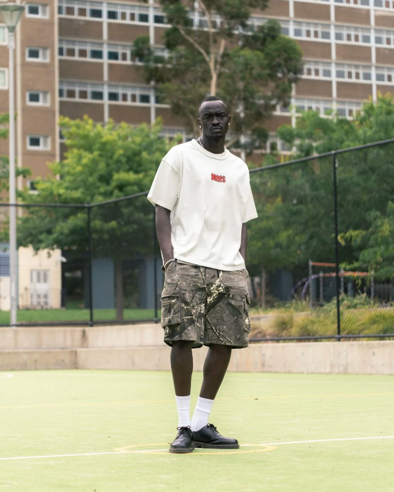 Person standing on a tennis court wearing a white t-shirt and camouflage shorts.