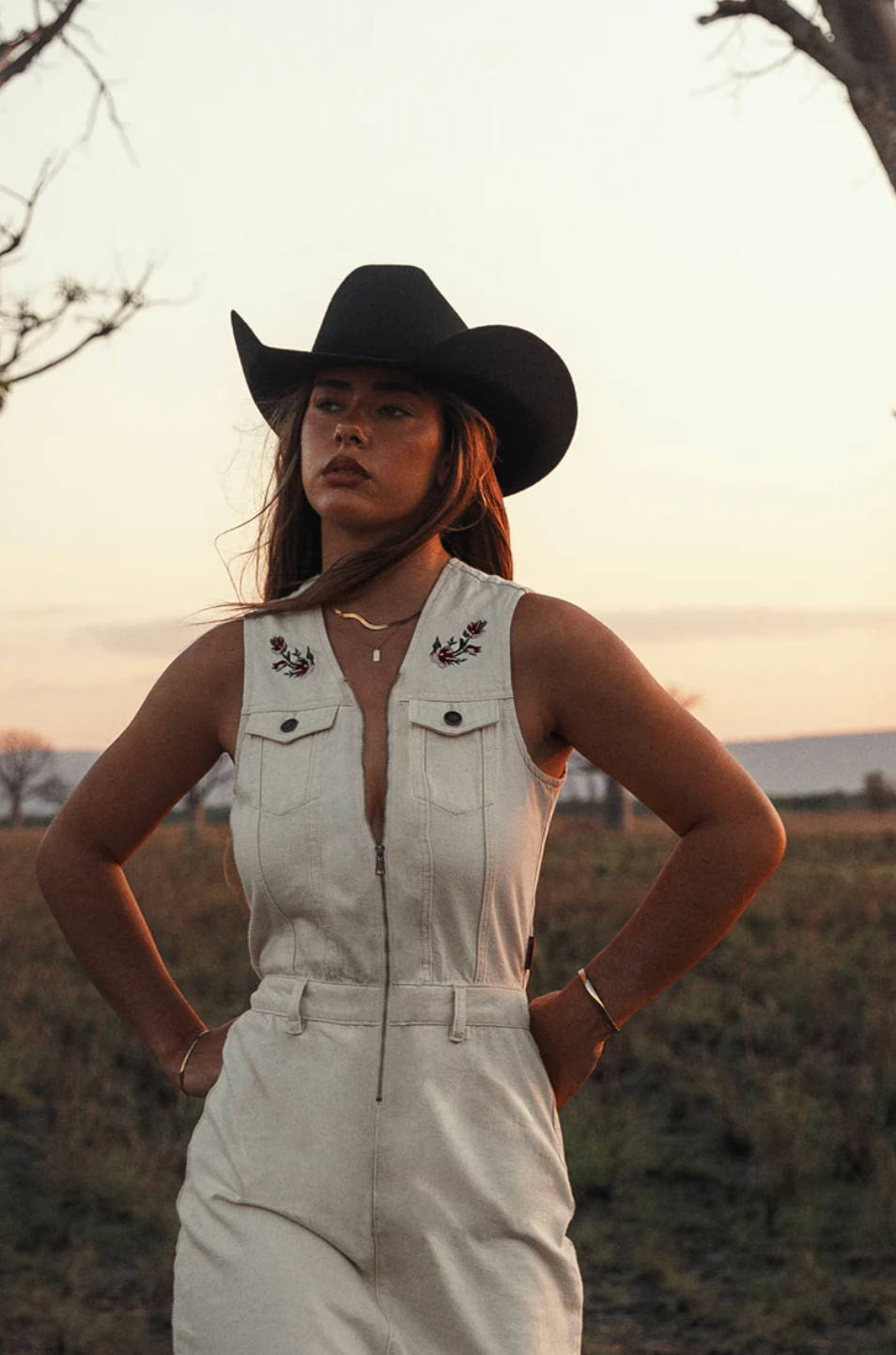 Woman wearing a white denim jumpsuit and black cowboy hat in an outdoor setting.