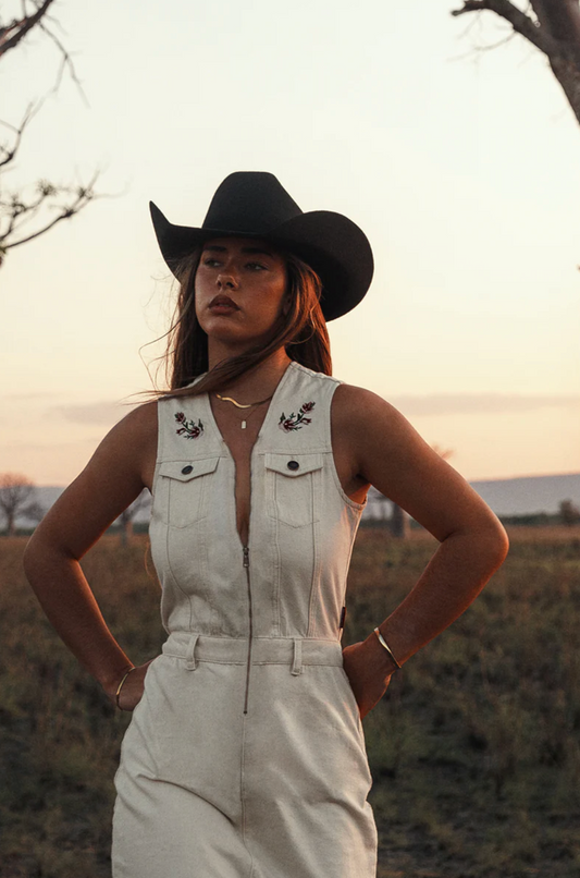 Woman wearing a white denim jumpsuit and black cowboy hat in an outdoor setting.