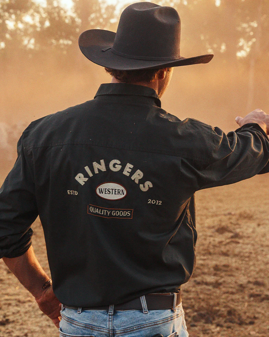 Person wearing a black 'Ringers' shirt with a cowboy hat and jeans, standing in a field.