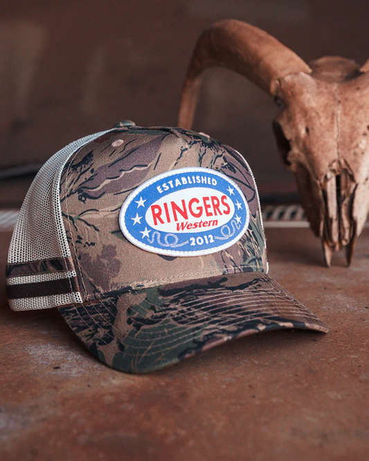 Camouflage cap with Ringers Western logo on a wooden surface with a skull in the background