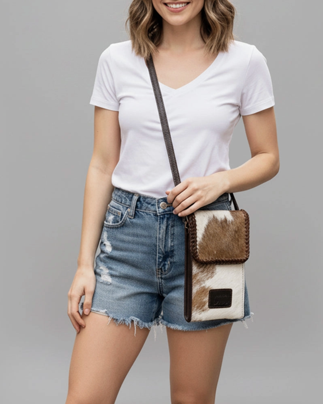 Woman wearing a white t-shirt and denim shorts, holding a cowhide crossbody bag on a gray background