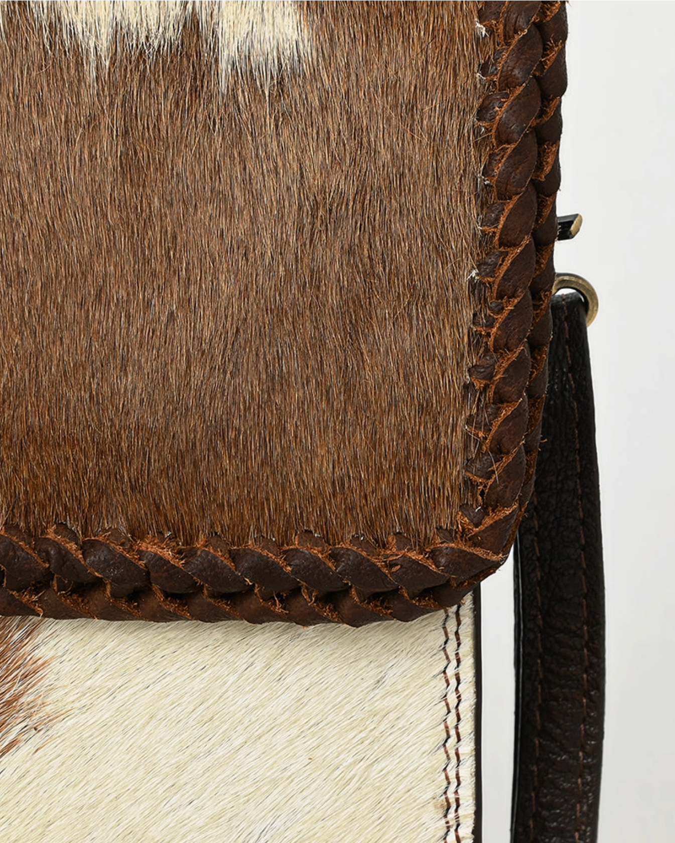 Close-up of a brown and white cowhide clutch with braided leather trim on a light gray background.